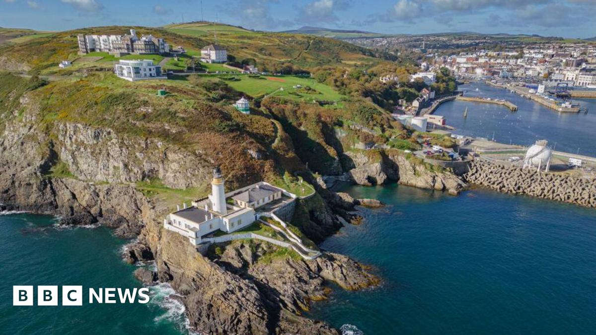 An arial view of Douglas Head, you can see the lighthouse on a rocky terrain, there are buildings scattered on the green terrain.