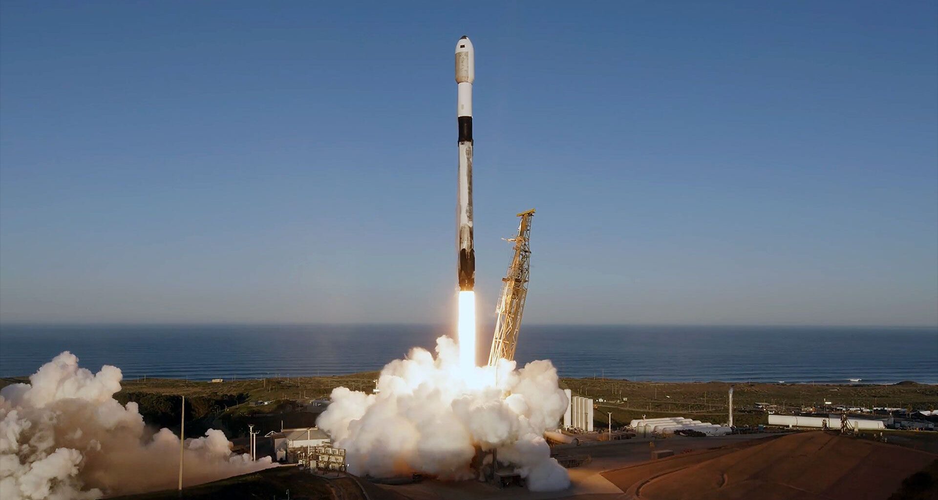 A white and black rocket launches into a clear blue sky backdropped by a large body of water