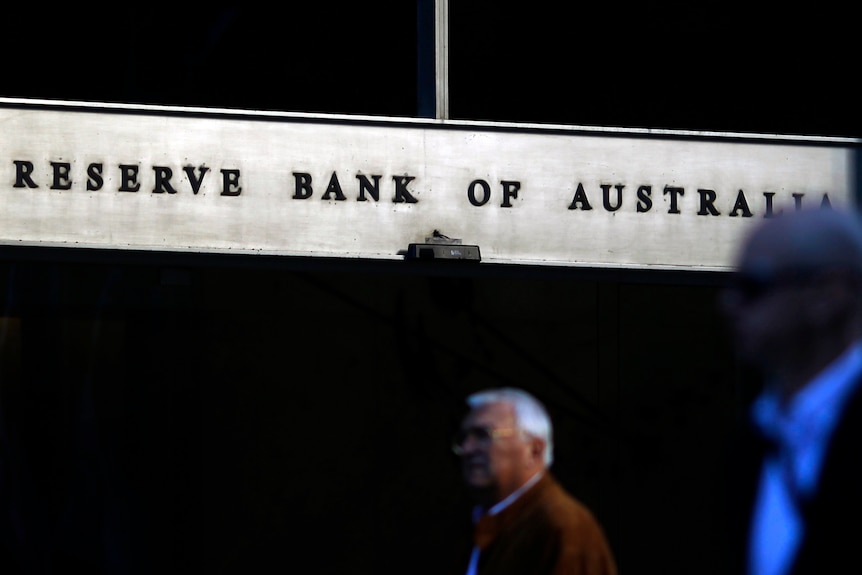 Two people walk past a steel sign reflecting in the sunlight that reads "Reserve Bank of Australia".