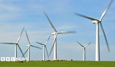 Six wind turbines with blades that are blurry, suggesting they are moving faster than the camera can capture them. A flock of sheep can be seen in the foreground on a green grass landscape.