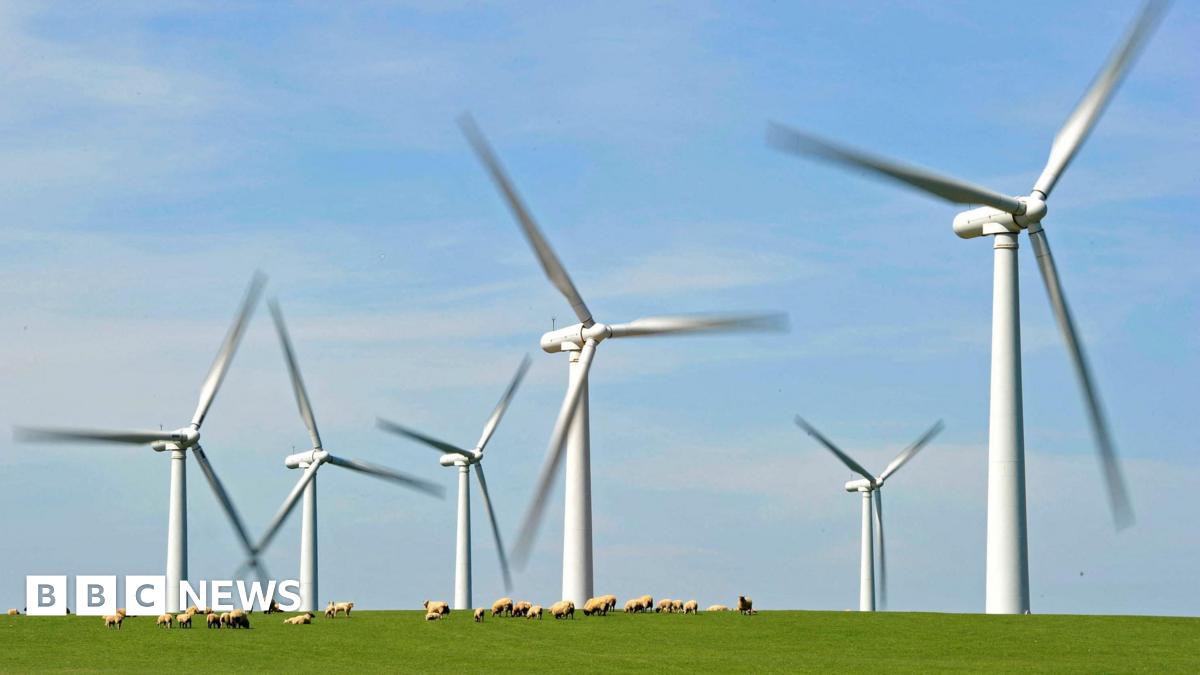 Six wind turbines with blades that are blurry, suggesting they are moving faster than the camera can capture them. A flock of sheep can be seen in the foreground on a green grass landscape.