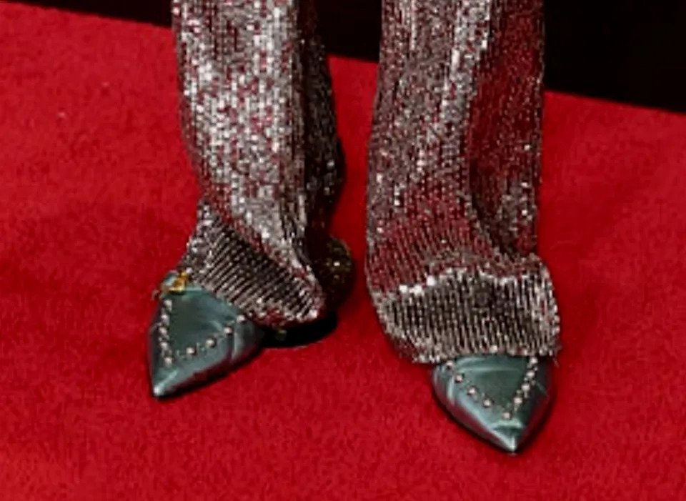 Person on red carpet wearing a shimmering, patterned suit with a sheer top. Grammy awards backdrop with golden gramophone trophies visible