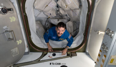 A man with dark hair and a mustache looks up at the camera, smiling. His hands are on the bottom edge of the hatch entrance as he makes his way inside the space station. The path he is coming through has many square cargo bags strapped to each side.