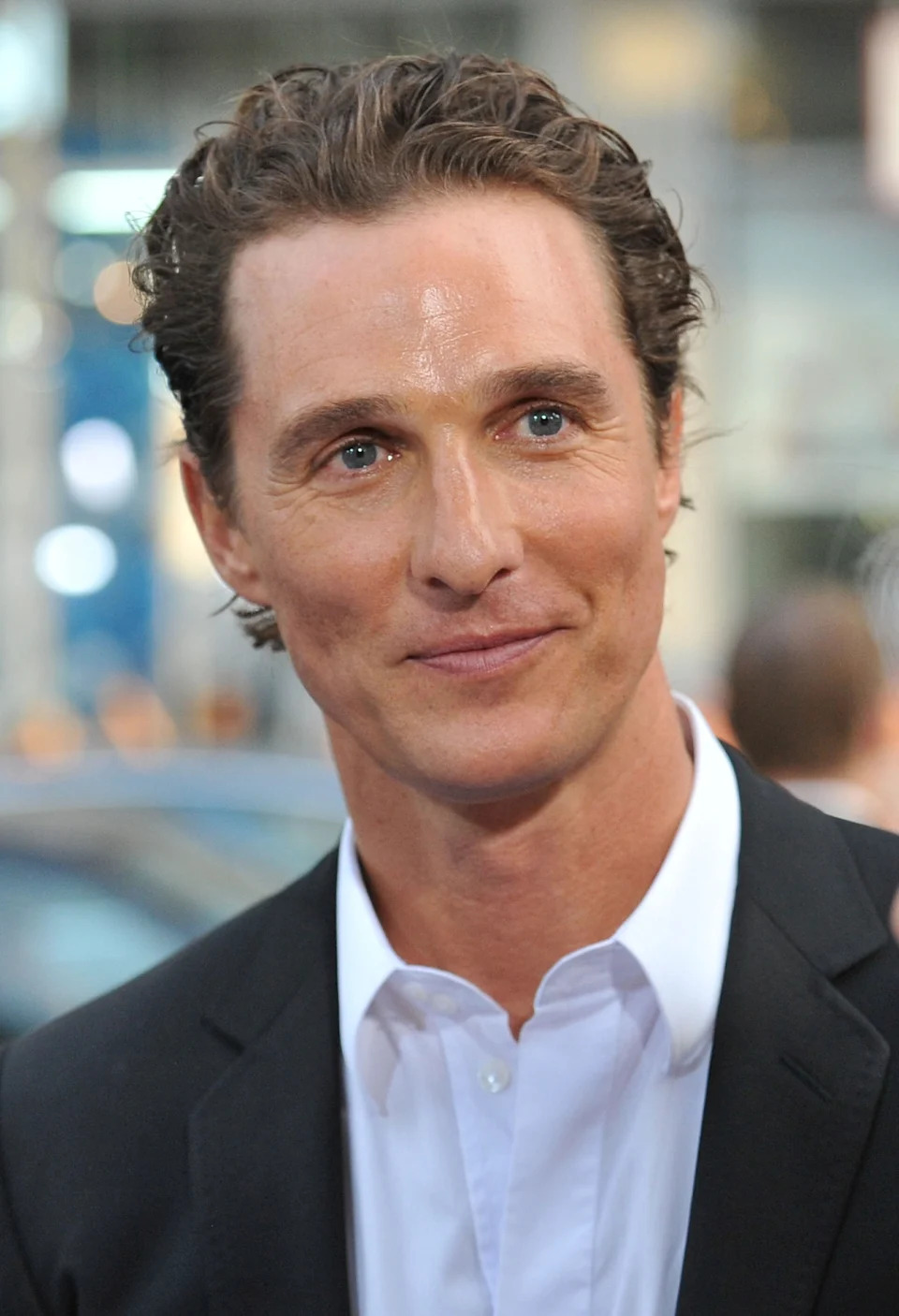 A man in a smart suit with a white shirt stands on a red carpet, looking ahead with a slight smile at a formal event