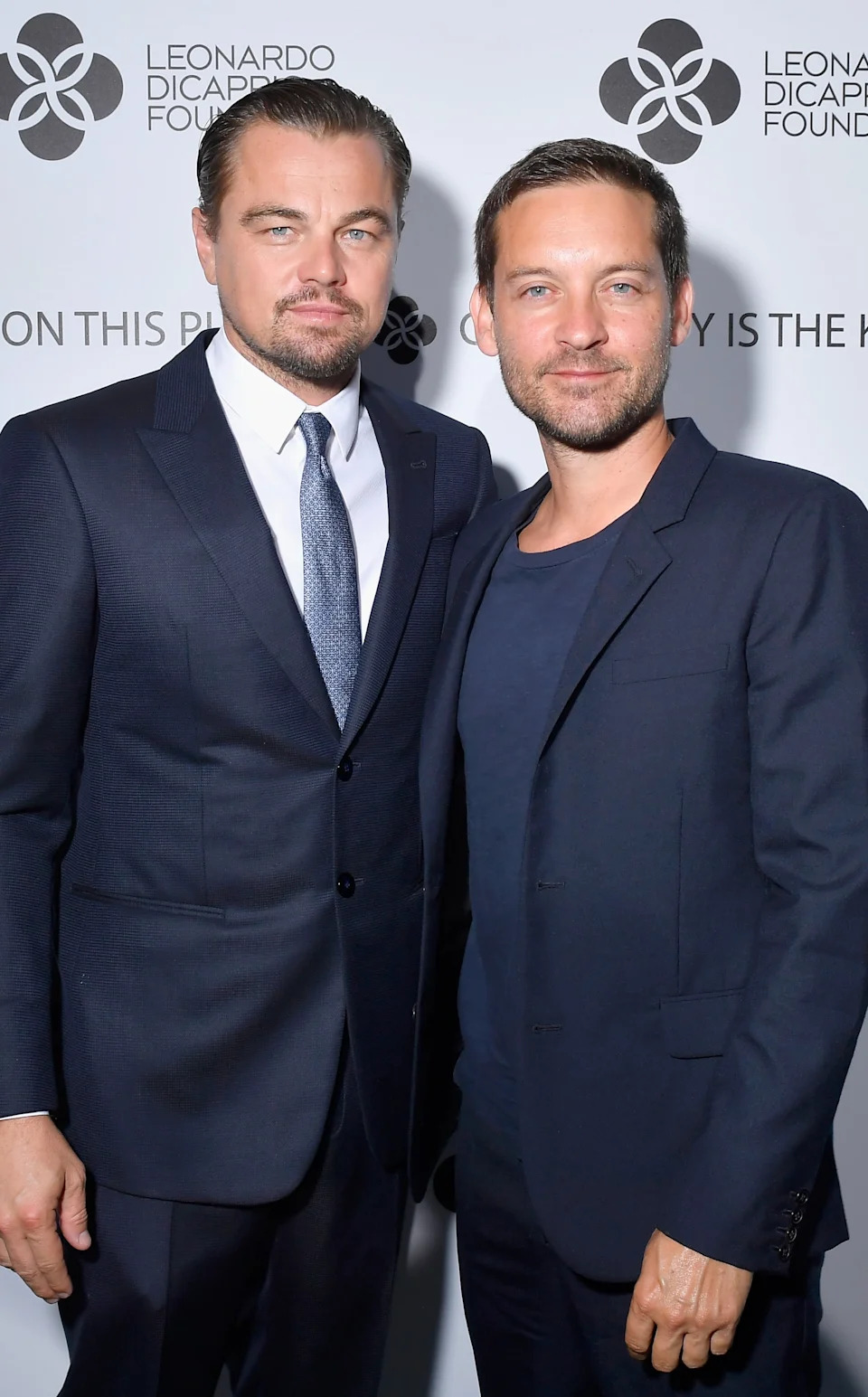 Two individuals at a Leonardo DiCaprio Foundation event, both in dark suits, posing for a photo against a branded backdrop