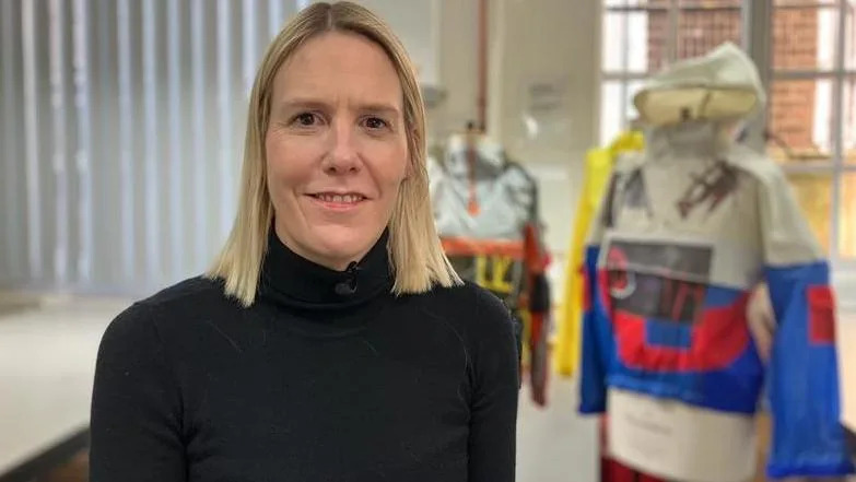 Sarah Graham has blonde shoulder-length hair and wears a black turtleneck top. Behind her are mannequins dressed in colourful garments made from boat sails featuring bold blocks of red, blue, yellow, and white.