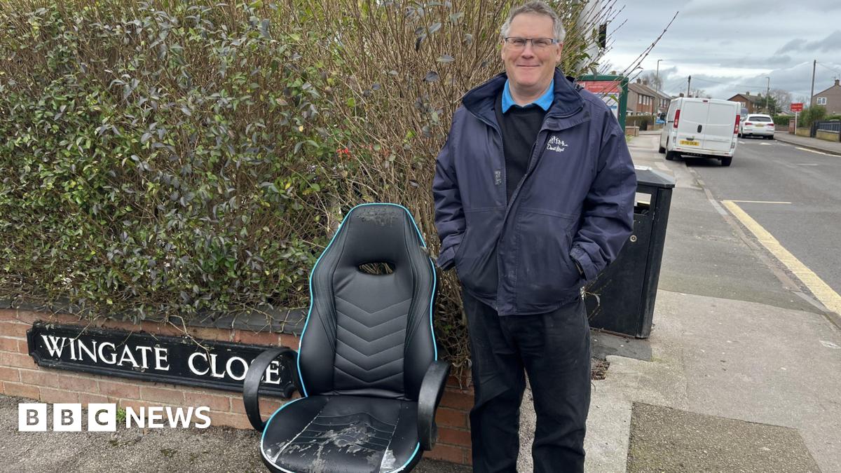 A man with grey hair and a navy blue coat stands at the end of a residential street corner, signed 'Wingate Close'. He is stood with his hands in his pockets next to a worn down black leather office chair on the pavement.