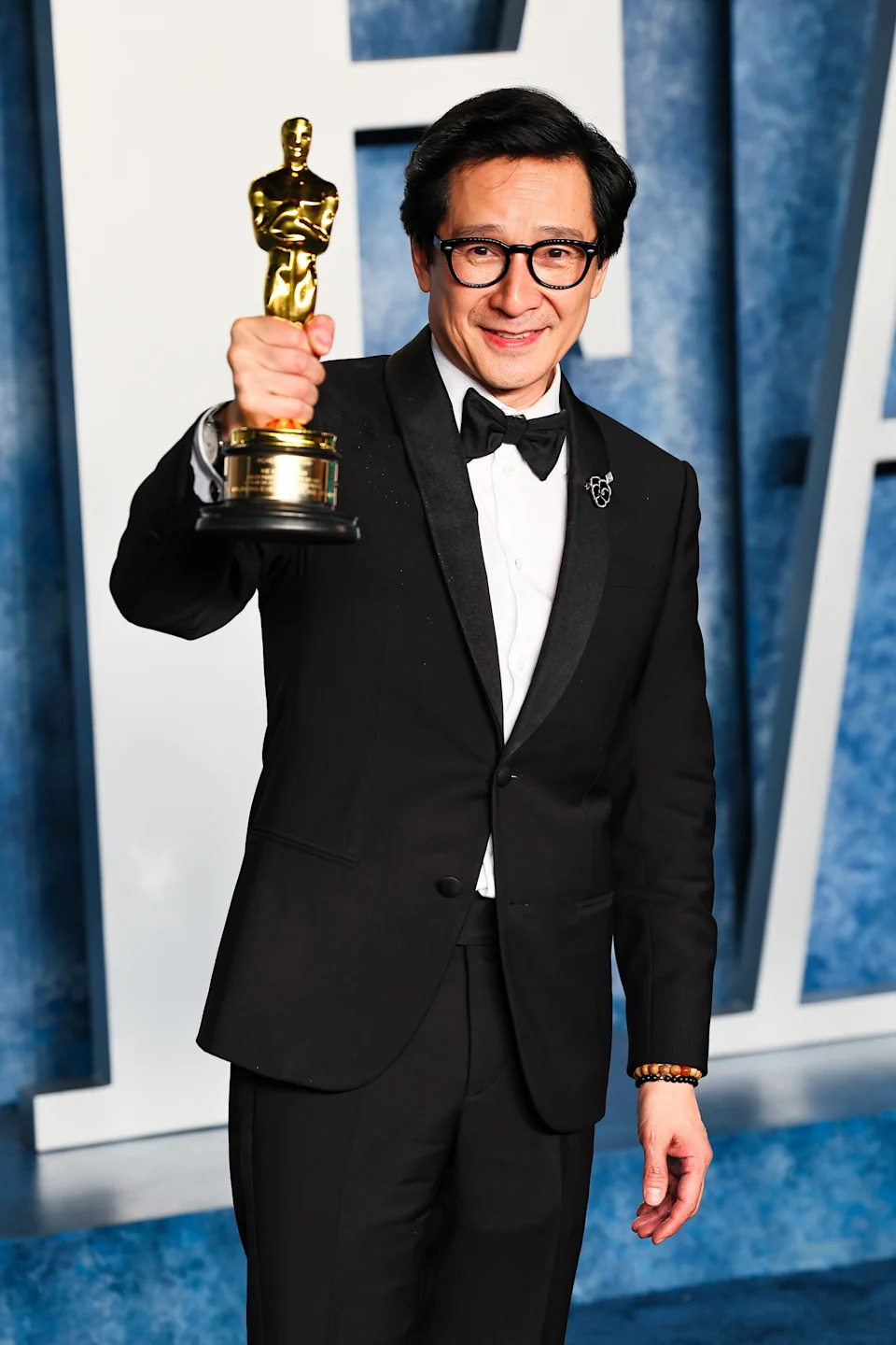 Person in a tuxedo holding an award statue, smiling, standing on what appears to be an event backdrop