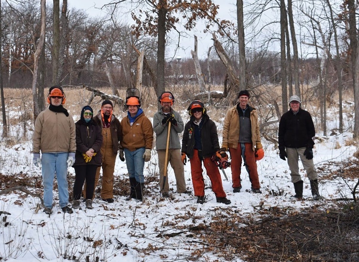 A group of seven people wearing winter clothing and safety gear stand in a snowy forested area, holding tools and equipment.
