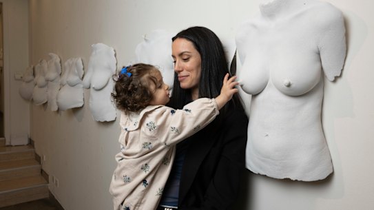 Amelie Cazzulino’s breastfeeding daughter, Gabrielle reaches towards a cast of her mother in the exhibition The Wall of Wombs by artist Juliana Monteiro.