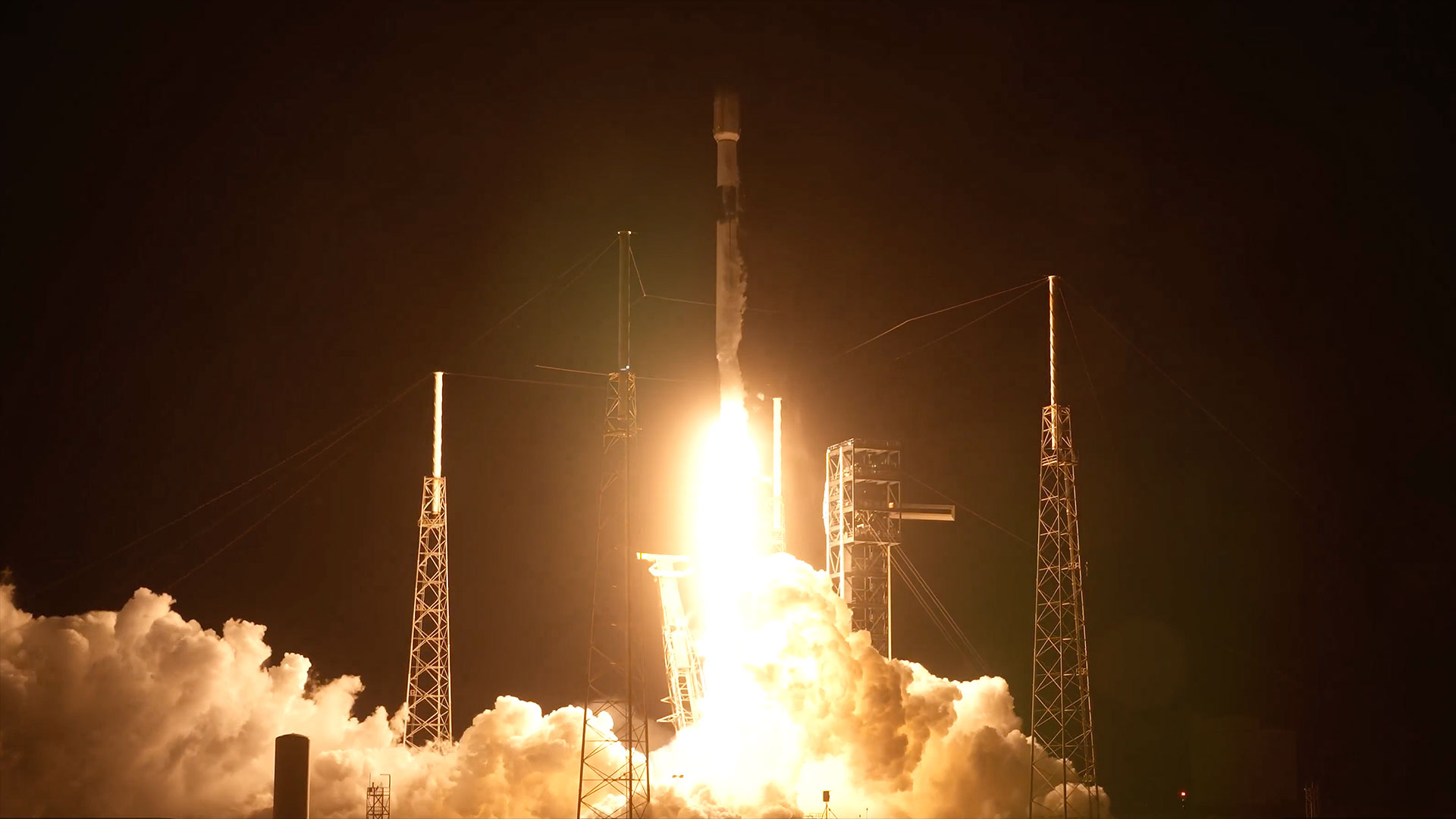 a white and black rocket lifts off into the night sky as its orange-white engine plume lights its launch pad below.
