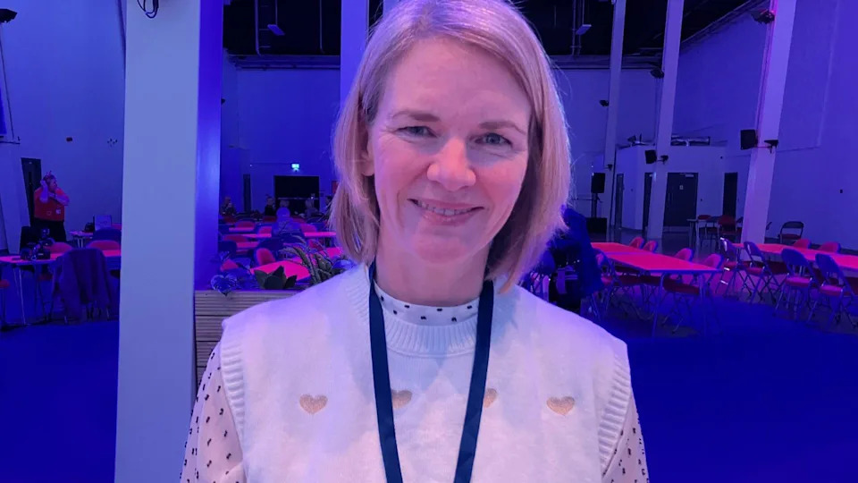 A woman with blonde bobbed hair wearing a white blouse and a cream tank top with hearts on. She is smiling and wearing a blue lanyard. There are tables and chairs in the background. 