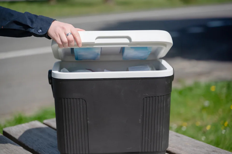 A hand opens a cooler on a picnic table, revealing stored items inside