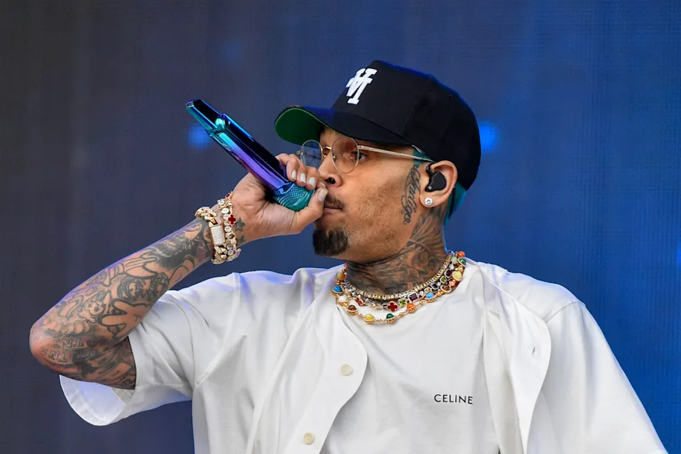 Chris Brown wearing a baseball cap, glasses, and a shirt sings into a microphone on stage