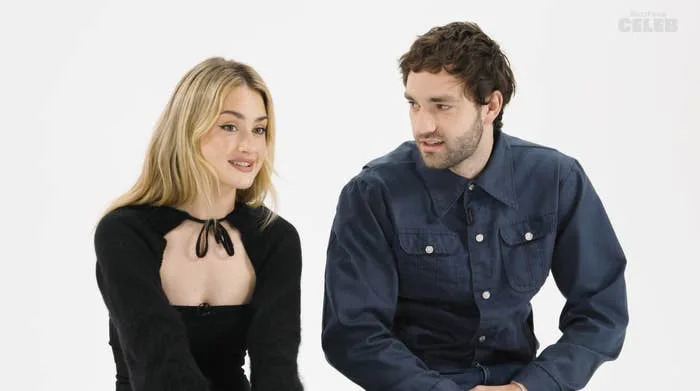 Two individuals sit together. The woman wears a black outfit with a bow detail. The man is in a denim shirt. Both look at each other, smiling