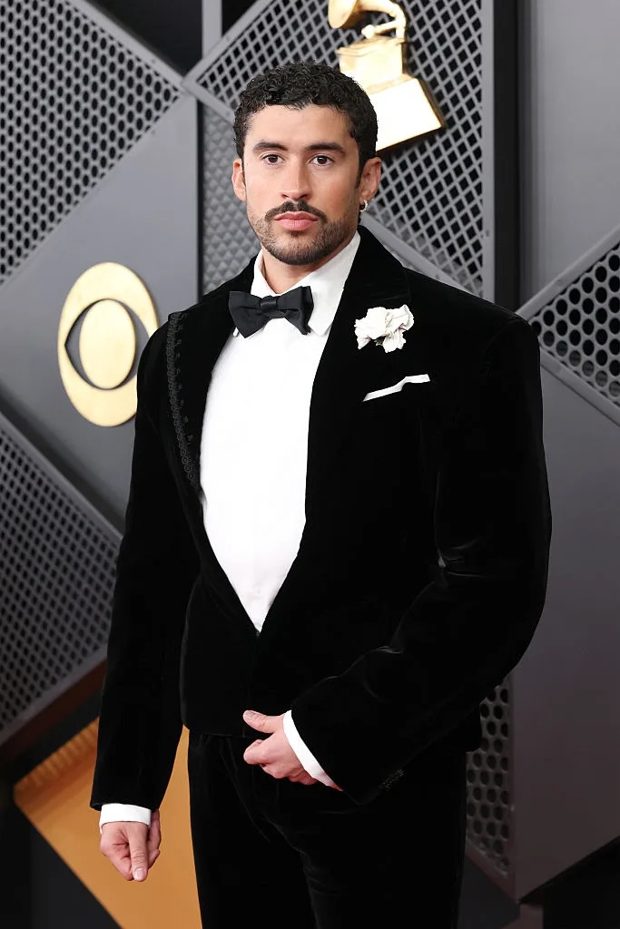 Person in velvet tuxedo with a bow tie and floral boutonniere at a formal event. Geometric backdrop with a Grammy trophy in the background