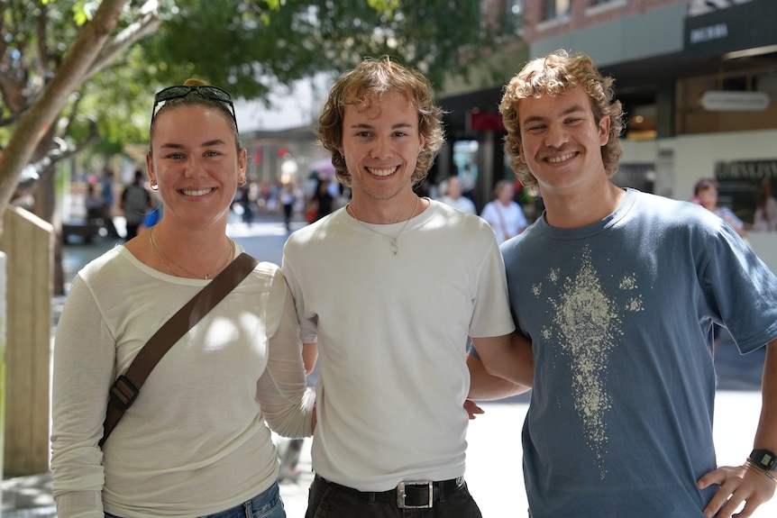 Three young people, a woman and two men, stand together smiling outside.