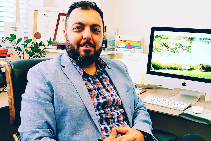 A man in a checked shirt and suit jacket and pants sitting at a desk chair smiling for the camera