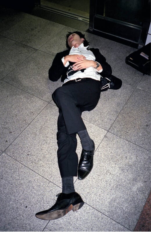 A person in a suit lies on the ground indoors with their eyes closed, hands resting on their chest, and legs crossed, appearing to be asleep or resting.