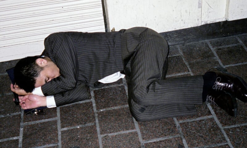 A man in a pinstripe suit lies curled up on a tiled sidewalk, resting his head on his arm, appearing to be asleep.