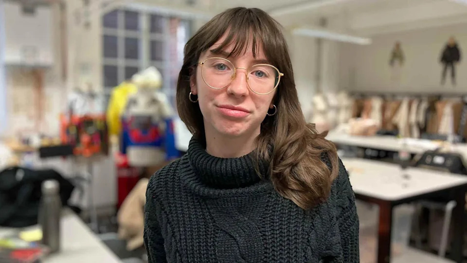 Ruby Ormston is wearing a dark knitted sweater and has shoulder‑length wavy hair and gold-rimmed glasses. In the background, the studio is filled with large tables, sewing equipment, and mannequins draped in colorful material. 