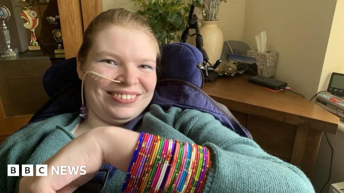 Lucinda Ritchie is smiling, seated in a supportive chair raising an arm covered with multiple colourful beaded bangles. She has an oxygen tube. She is wearing a textured, long‑sleeved top. In the background are a wooden cabinet with trophies, a desk with assorted items, and decorative plants.