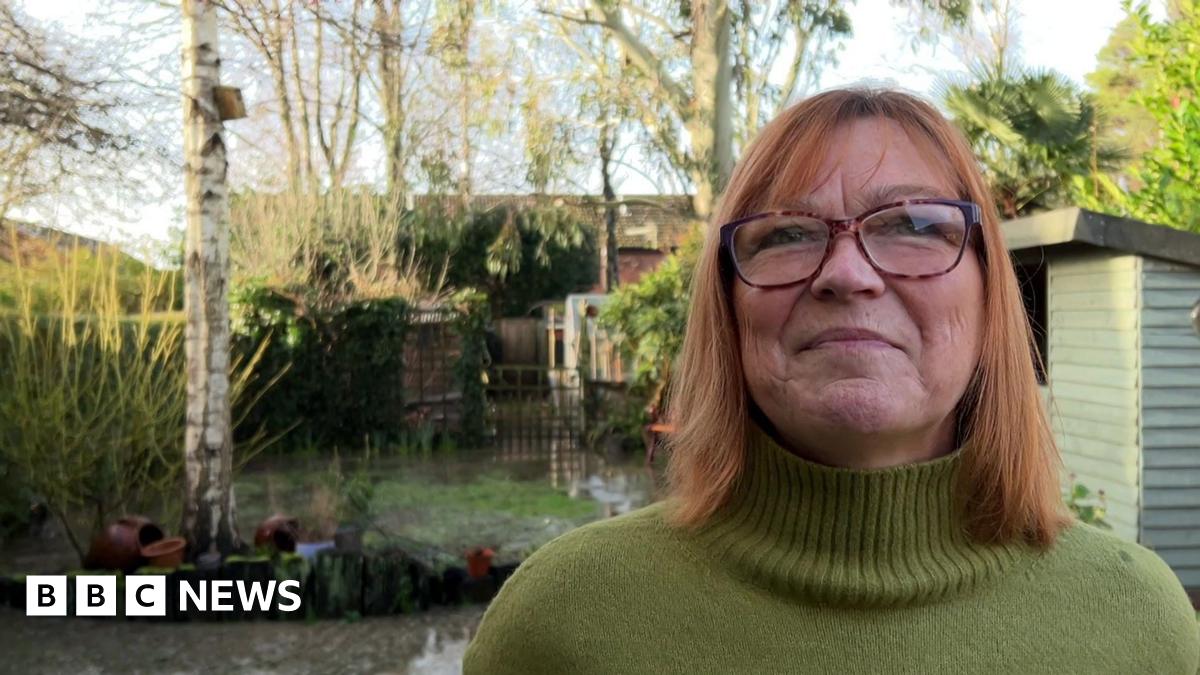 Susannah Gill, who is wearing glasses and a green turtle-neck jumper, stands in her sewage-flooded garden