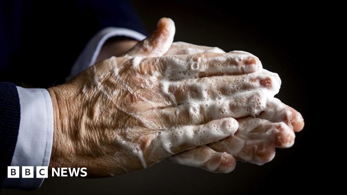 A close up image of the hands of a person who is wearing a suit. The picture has been taken while they are washing their hands and there is soap residue which is yet to be washed off.