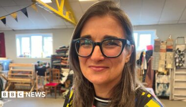 Melissa Santiago-Val is sat in an open spaced room. She has long brown hair and is wearing black frames glasses and a patterned top. Behind her are art supplies and a silver ladder.