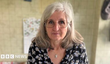 A woman with grey hair, glasses and a dark blue blouse with flowers is looking into the camera. She is standing in her kitchen.
