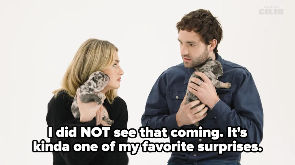 Two individuals seated, holding puppies, gaze at each other. One wears a cozy black outfit, the other a casual denim jacket