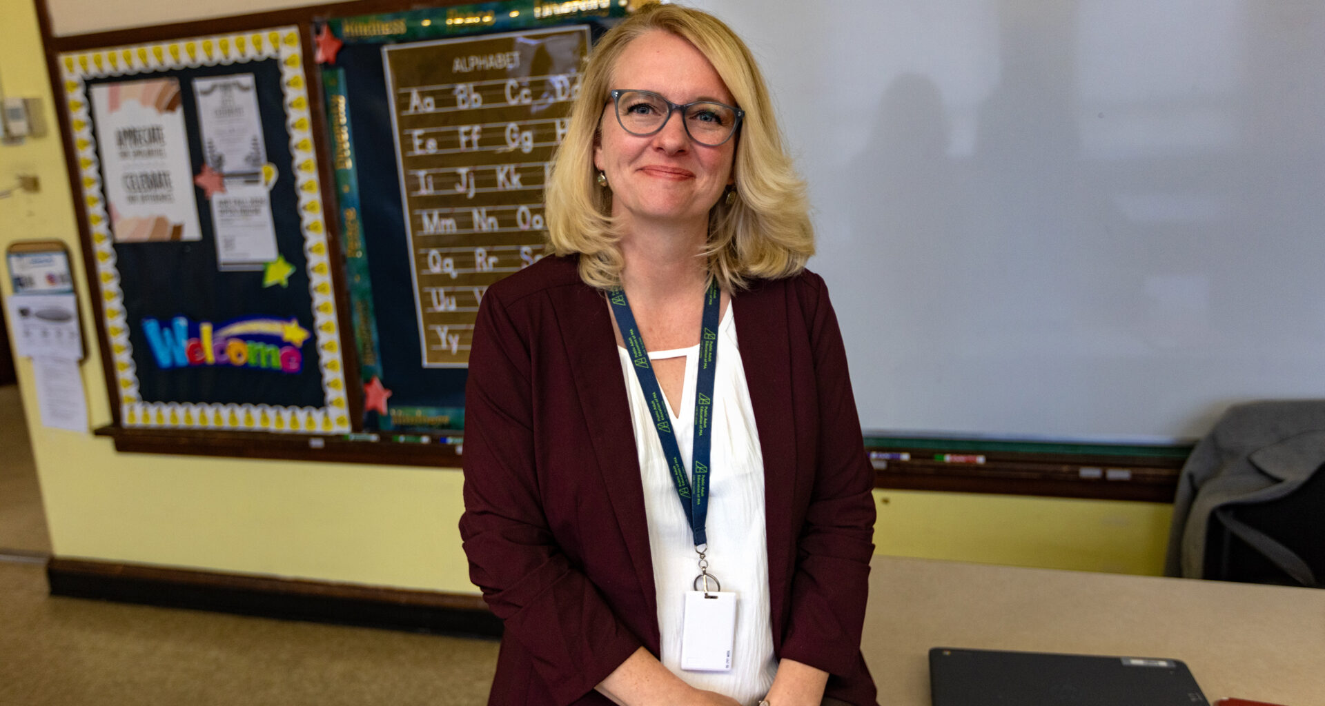 A blonde woman wearing glasses, a white shirt and suit with a lanyard around her neck.
