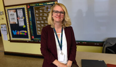 A blonde woman wearing glasses, a white shirt and suit with a lanyard around her neck.