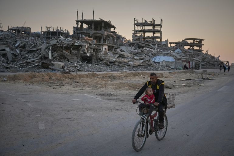 A man carries a child while riding his bicycle along a damaged street in the Al-Shati camp in Gaza City on November 18, 2025.