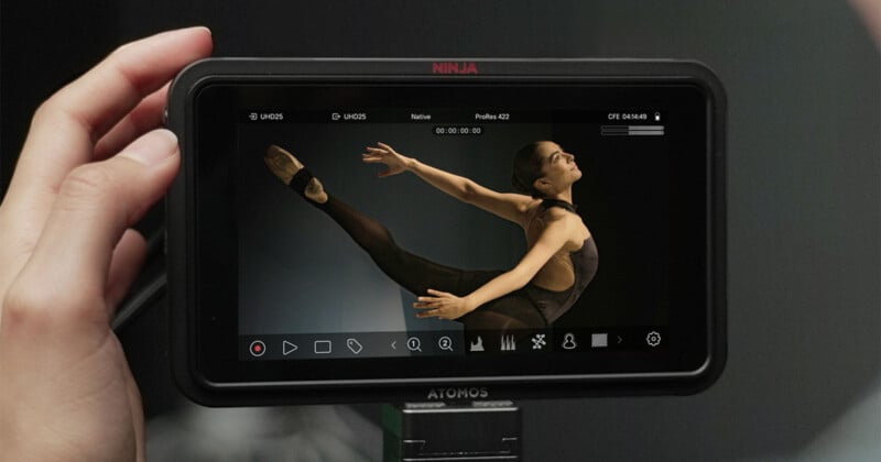 A hand holds a monitor displaying a video of a female ballet dancer in mid-air, performing an elegant leap with arms and legs extended gracefully against a dark background.