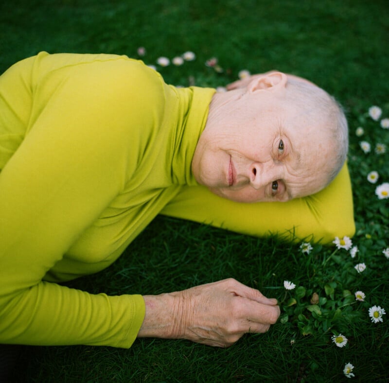 An older person with very short gray hair, wearing a bright yellow long-sleeve top, lies on green grass with small white flowers, resting their head on one arm and looking thoughtfully at the camera.