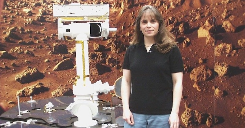 Woman with long hair and black shirt standing next to a robotic rover, in front of closeup image of rocky terrain.