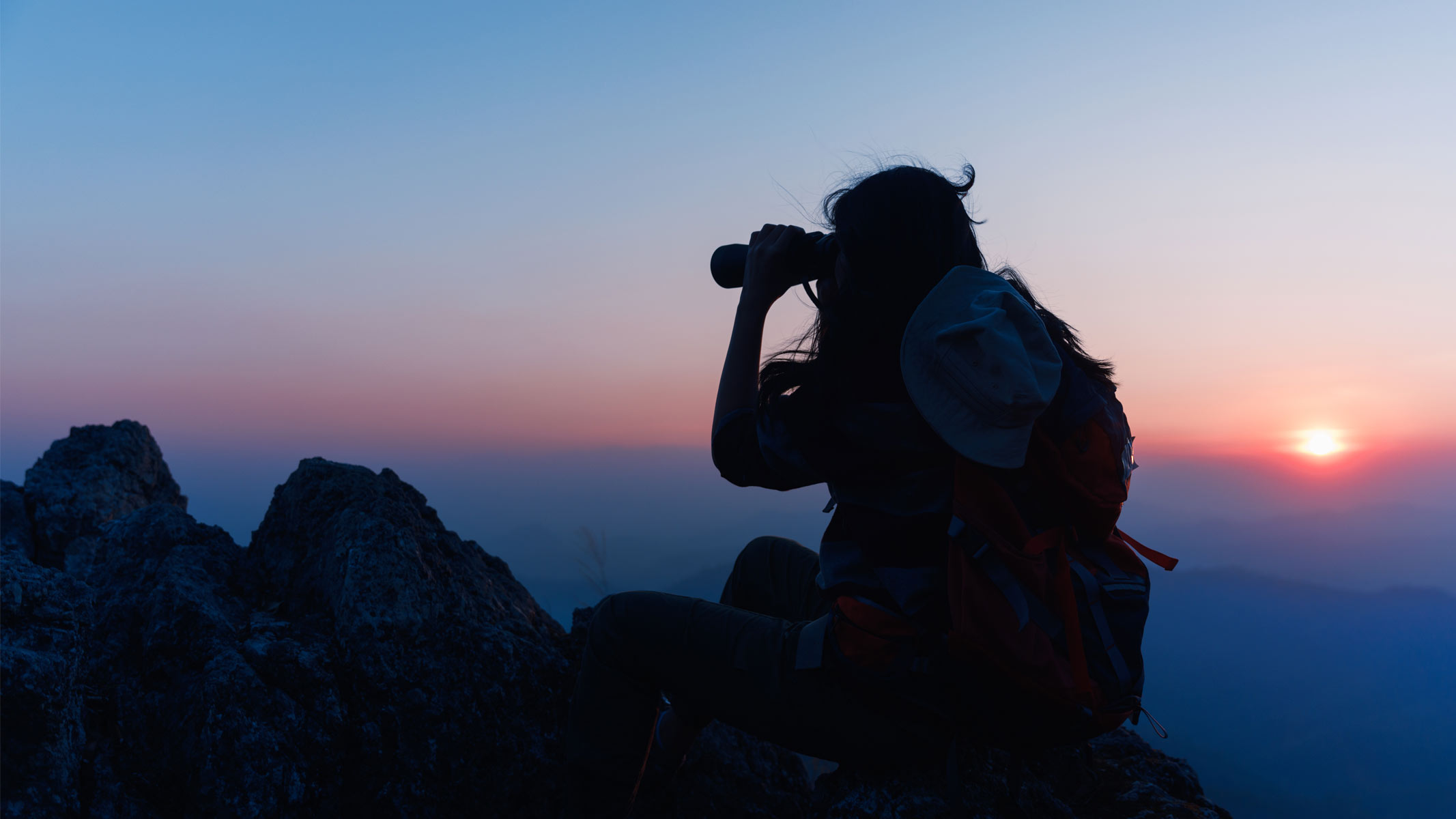 A silhouetted person using binoculars on top of a mountain with a twilight sky.