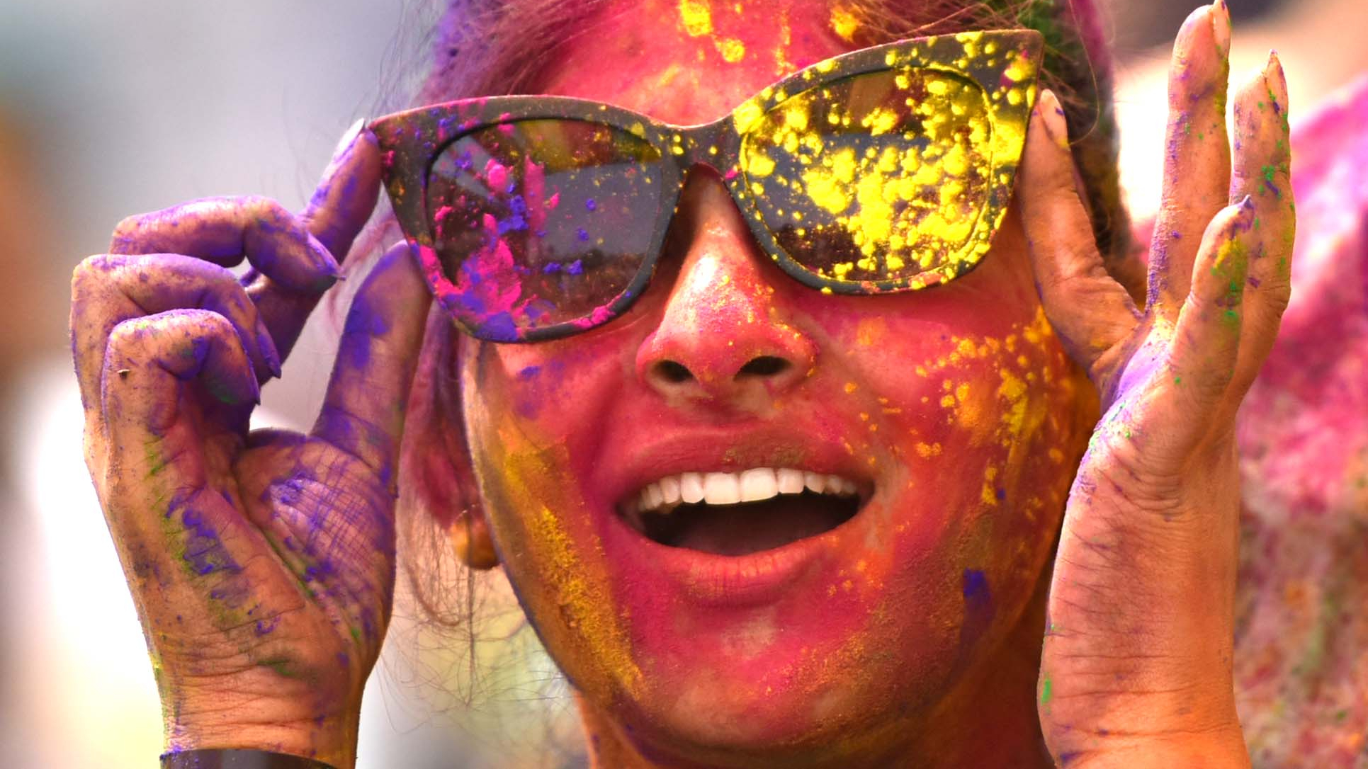A woman is pictured holding sunglasses to her face, while covered in bright, multi-colored paint and powder during the Holi Festival.