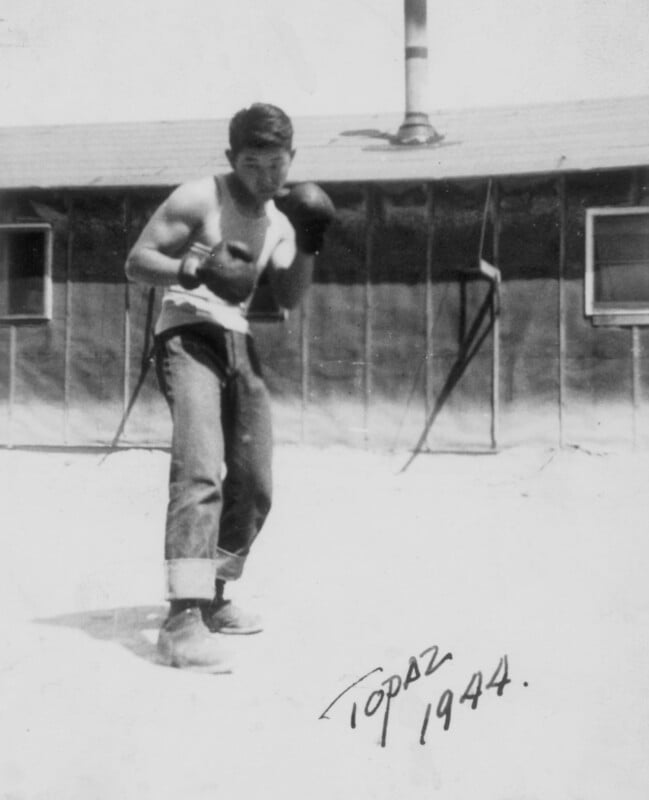 A young man wearing boxing gloves, a sleeveless shirt, cuffed jeans, and boots stands in a boxing stance outside a wooden building. The photo is labeled "Topaz 1944" in the bottom right corner.
