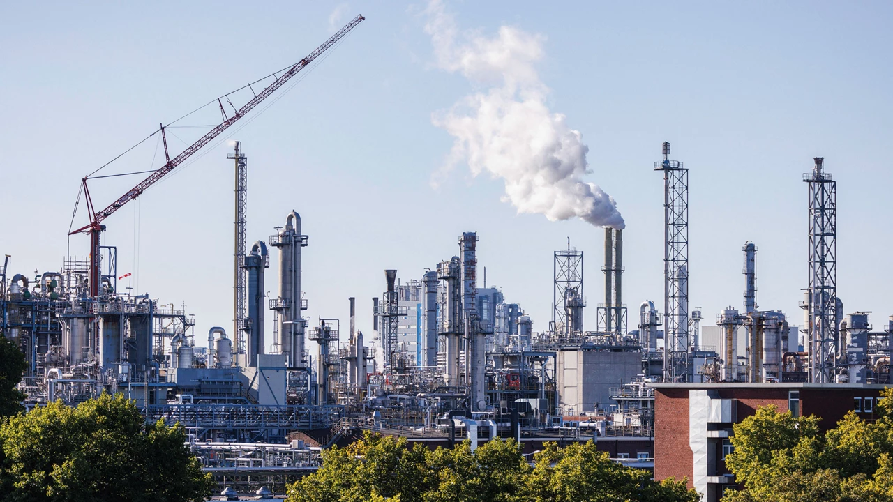 A large chemical plant with multiple buildings and metallic towers stands against a blue sky with trees in the foreground.
