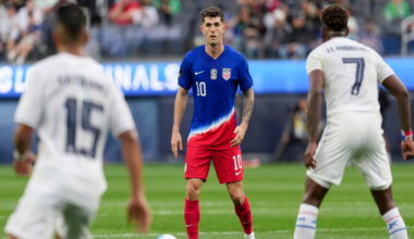 A soccer player in a blue and red US uniform stands poised with the ball, facing two opposing players in white uniforms.