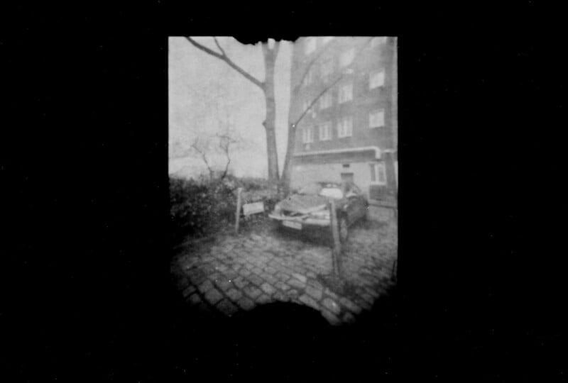 Black and white photo of an old, damaged car parked on a cobblestone street next to a tree and building, with a blurry, vignetted effect around the edges of the image.