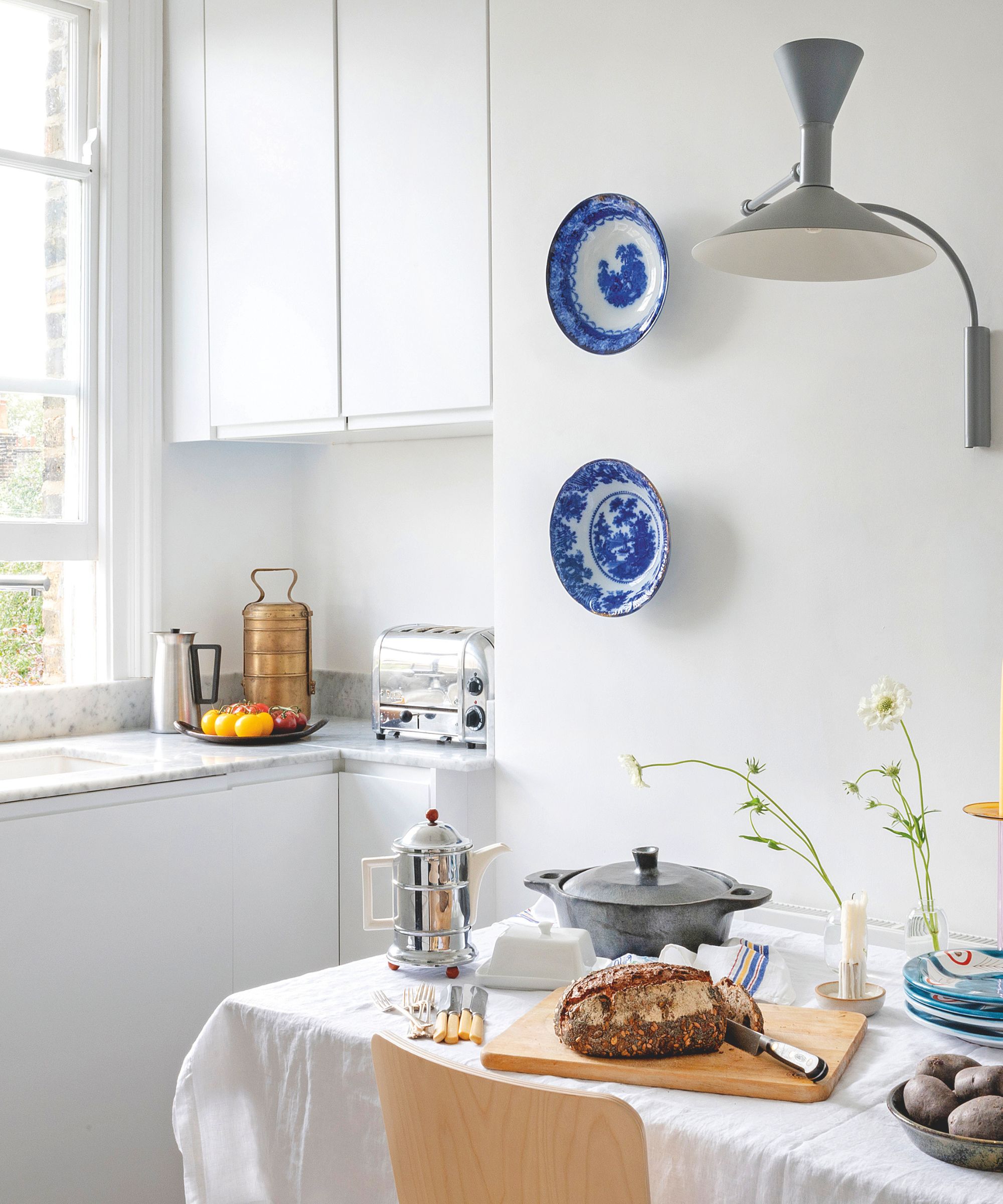 A bright, white kitchen with a marble countertop and a wooden table set for breakfast with fresh bread, a chrome teapot, and blue patterned plates hung on the wall