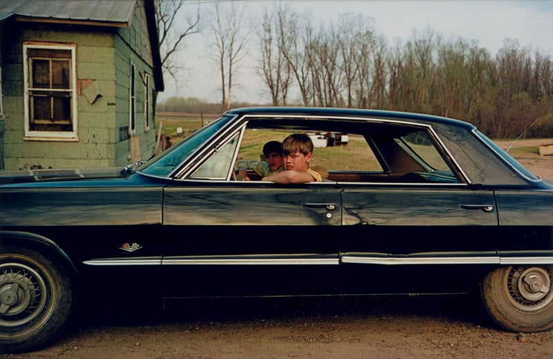 Two young boys sit in an old black car parked next to a weathered green house, with bare trees and open land visible in the background on a cloudy day.