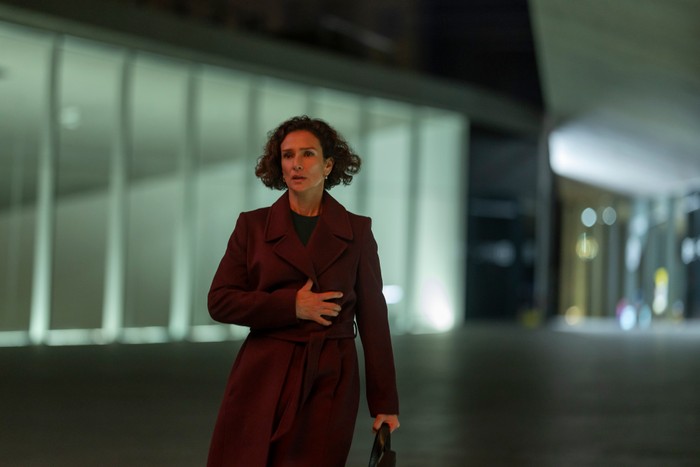 Indira Varma in The Night Manager season 2, wearing a red coat, stood outside at night and looking shocked.