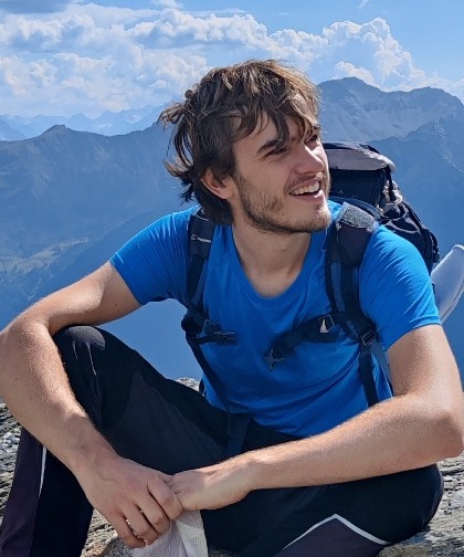 A young man with tousled hair and a beard, in hiking clothes and backpack, sits with mountains in the background.