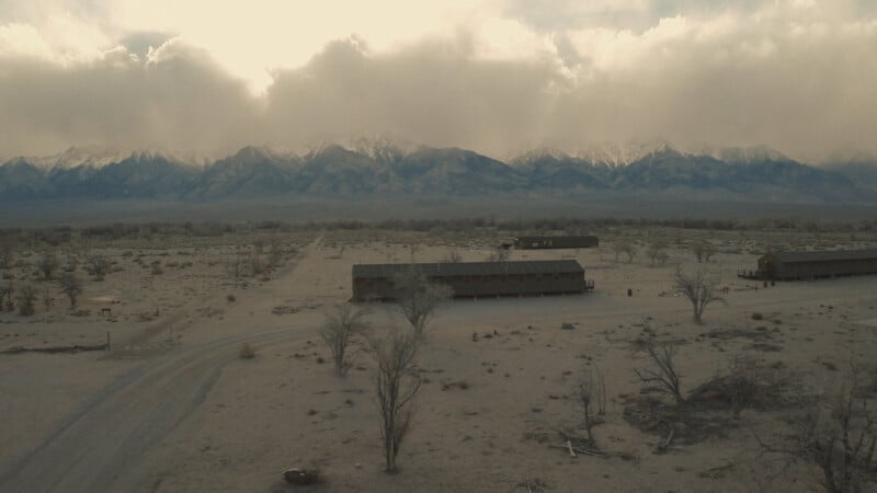 Desolate landscape with a few long, low buildings, leafless trees, and a dirt road; mountains with snow-capped peaks rise in the background under a cloudy sky.
