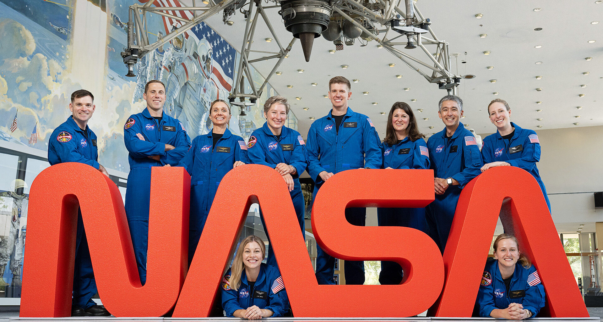 A group of NASA astronauts in blue flight suits pose together behind large red letters spelling “NASA.”