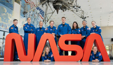 A group of NASA astronauts in blue flight suits pose together behind large red letters spelling “NASA.”